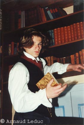 jeune homme romantique en chemise blanche et gilet noir devant une bibliothèque, un livre relié à la main